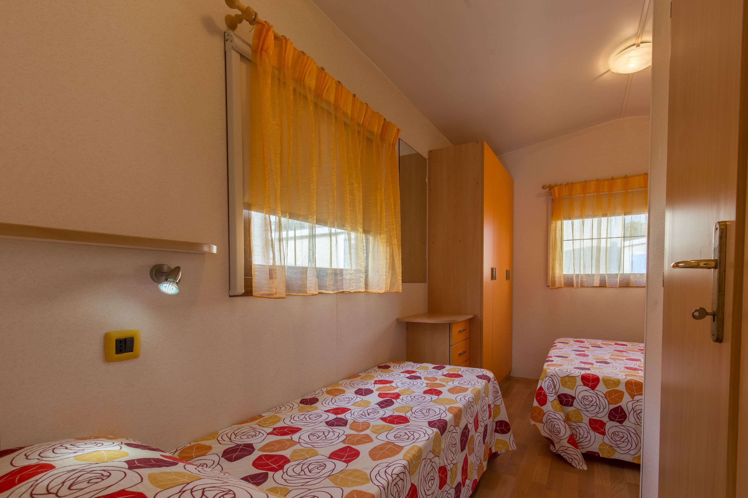 A small, well-lit room with two single beds, a mirror, a small table, and a window with curtains.