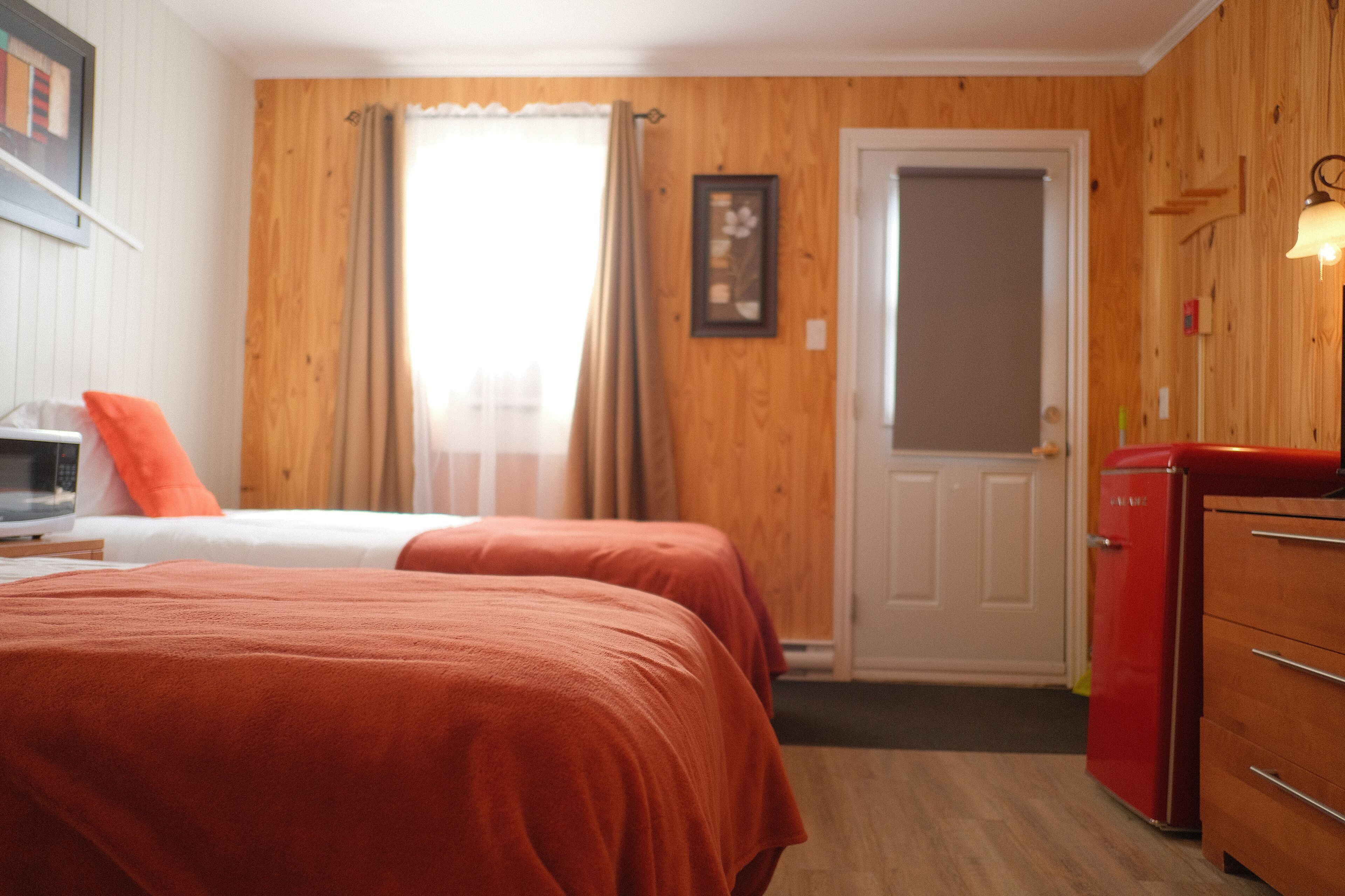 Ein Zimmer mit zwei Betten, einer Holzwand, einer Tür, einem roten Gerät und einem kleinen Tisch mit einer Mikrowelle.