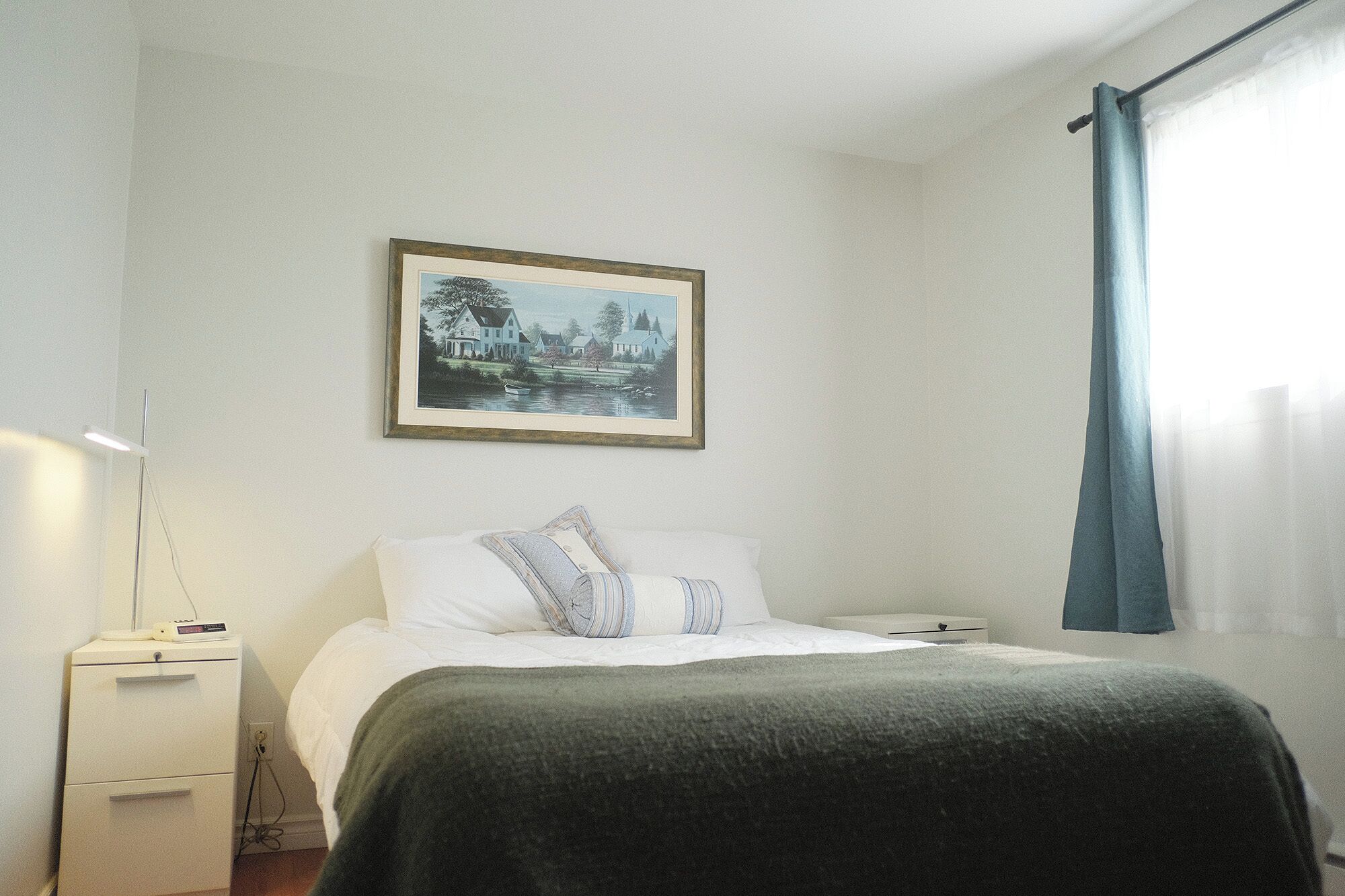 A bedroom with a bed, a bedside table, a lamp, a framed picture on the wall, and a window with curtains.