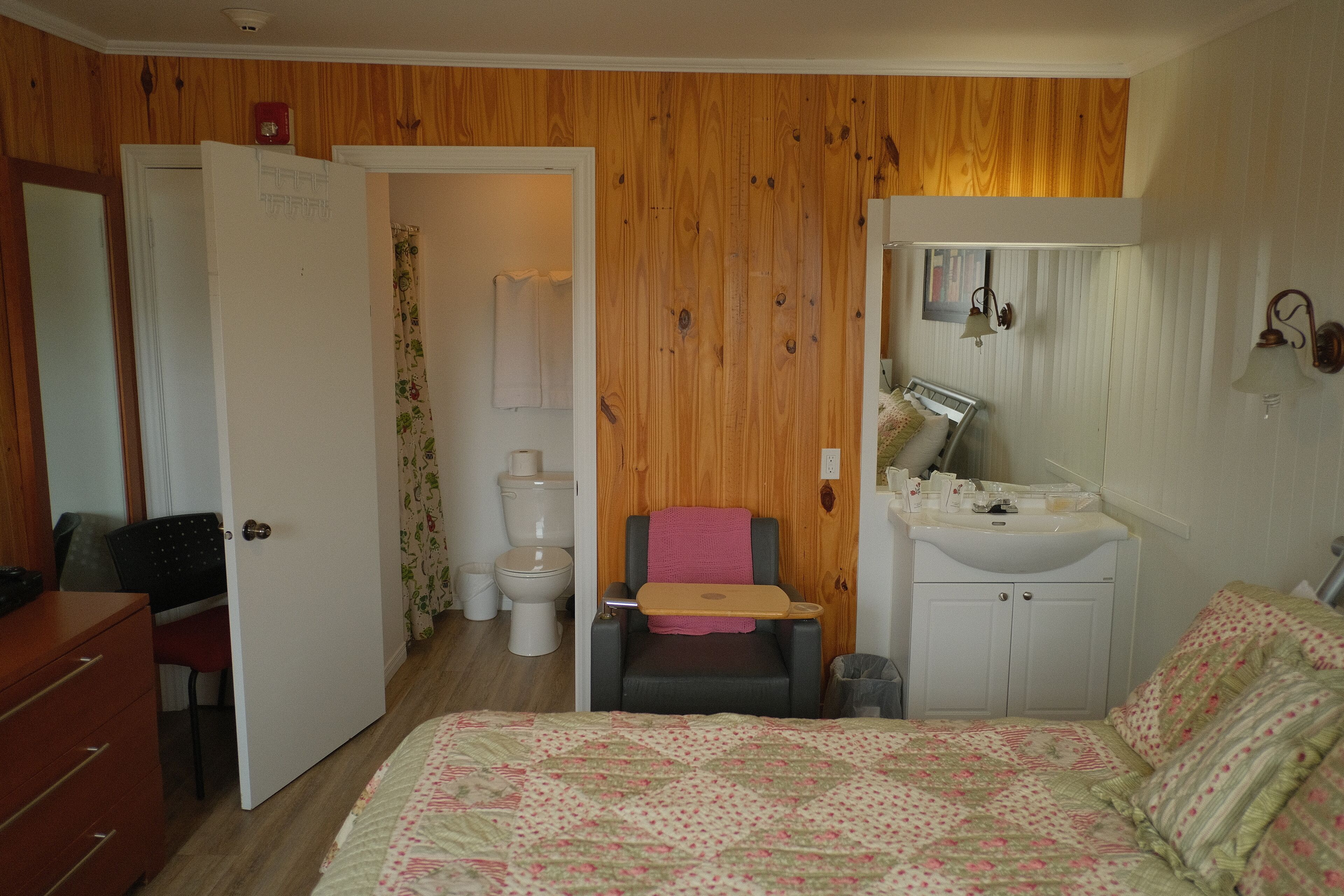 A bedroom with a bed, a desk, a chair, a small bathroom with a toilet, and a sink.