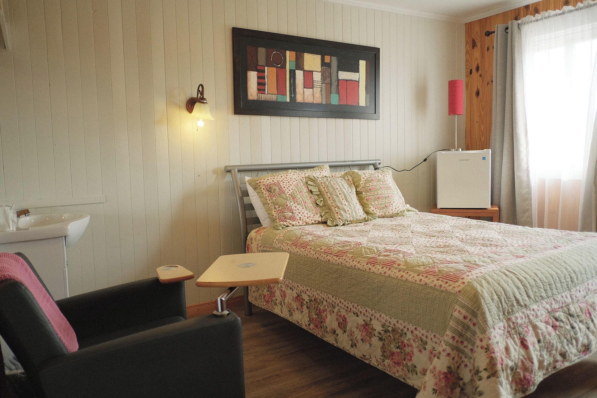 A bedroom with a bed, a nightstand, a chair, a desk, a wall-mounted lamp, a framed artwork, and a window with curtains.