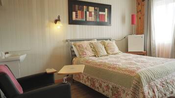 A bedroom with a bed, a nightstand, a chair, a desk, a wall-mounted lamp, a framed artwork, and a window with curtains.