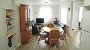 A dining area with a wooden table and wicker chairs, a television, and a sofa in the background. - Auberge St-Tite-des-Caps (Saint-Tite-des-Caps)