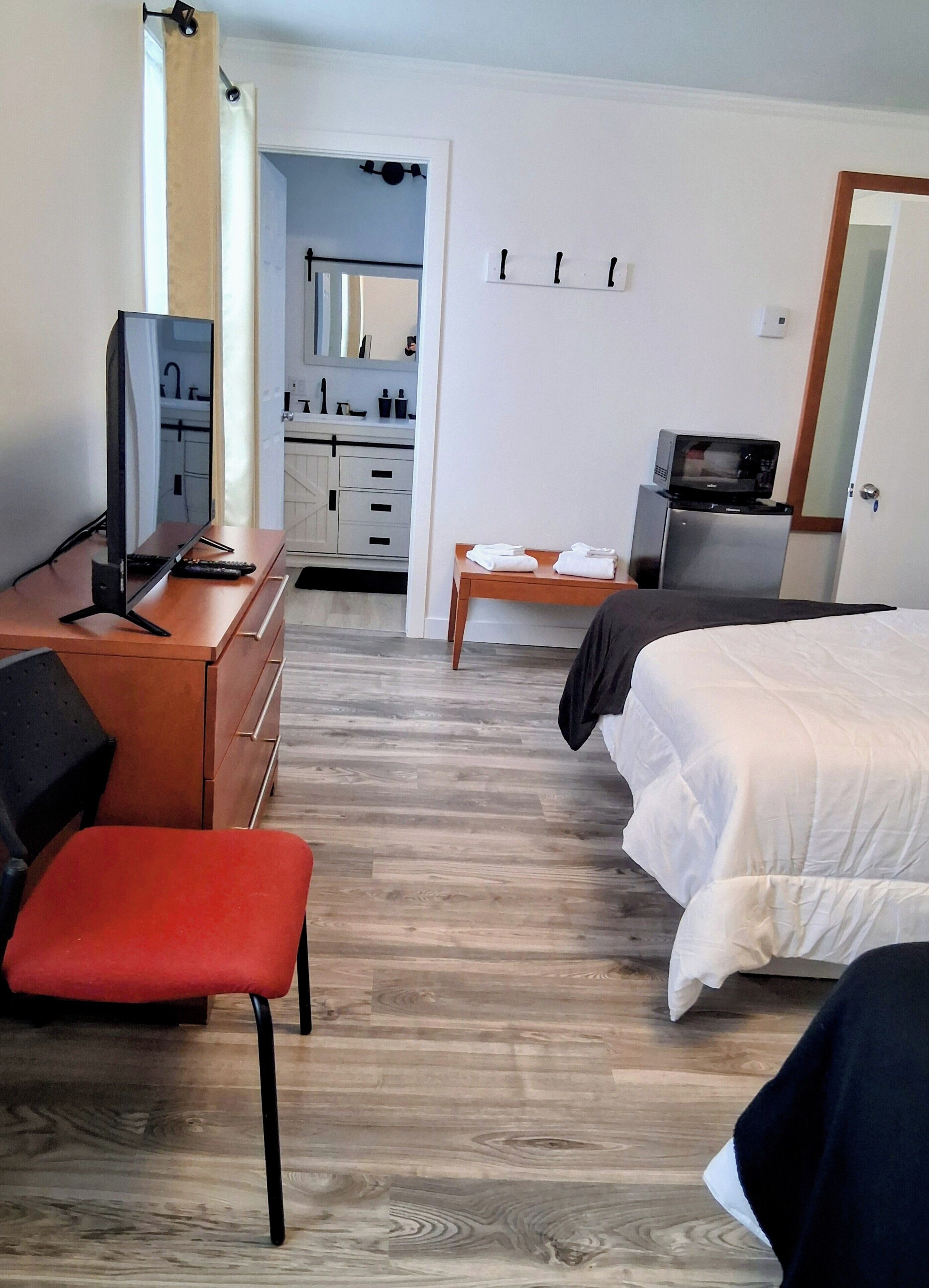 A bedroom with a bed, a desk with a computer, a chair, a small table with towels, and a microwave.