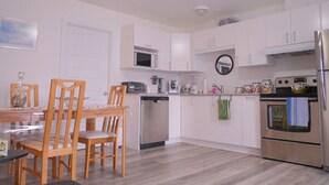 A modern kitchen with white cabinets, a dishwasher, a microwave, a refrigerator, and a dining table with chairs. - Auberge St-Tite-des-Caps (Saint-Tite-des-Caps)