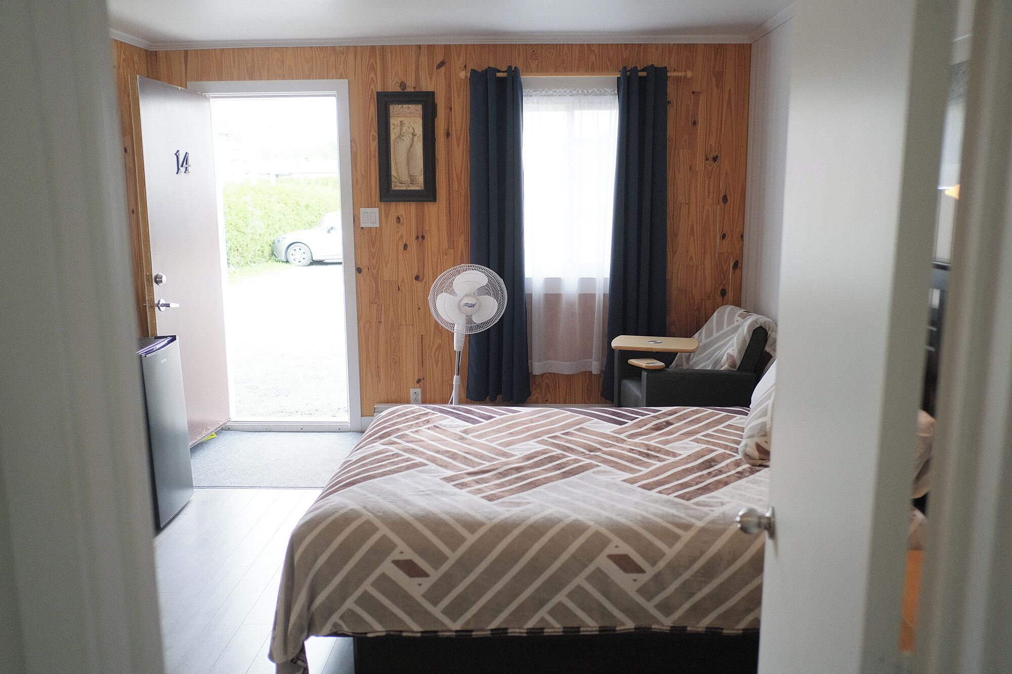 A hotel room with a bed, a desk, a chair, a standing fan, and a door leading to a parking area.