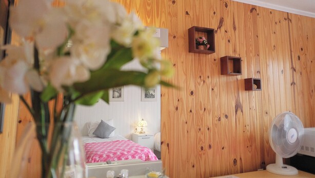 A bedroom with a wooden wall, a bed with a pink and white bedspread, a bedside table with a lamp, and a small wooden shelf with a picture. - Auberge St-Tite-des-Caps (Saint-Tite-des-Caps)