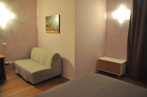 A small, neatly arranged bedroom with a single bed, a sofa, a small cabinet, and a painting on the wall.
