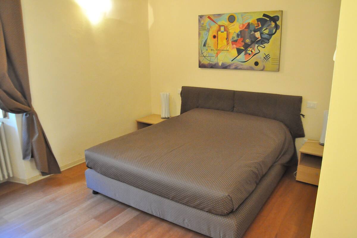 Een slaapkamer met een bed, nachtkastje, lamp, schilderij aan de muur en een raam met gordijnen.