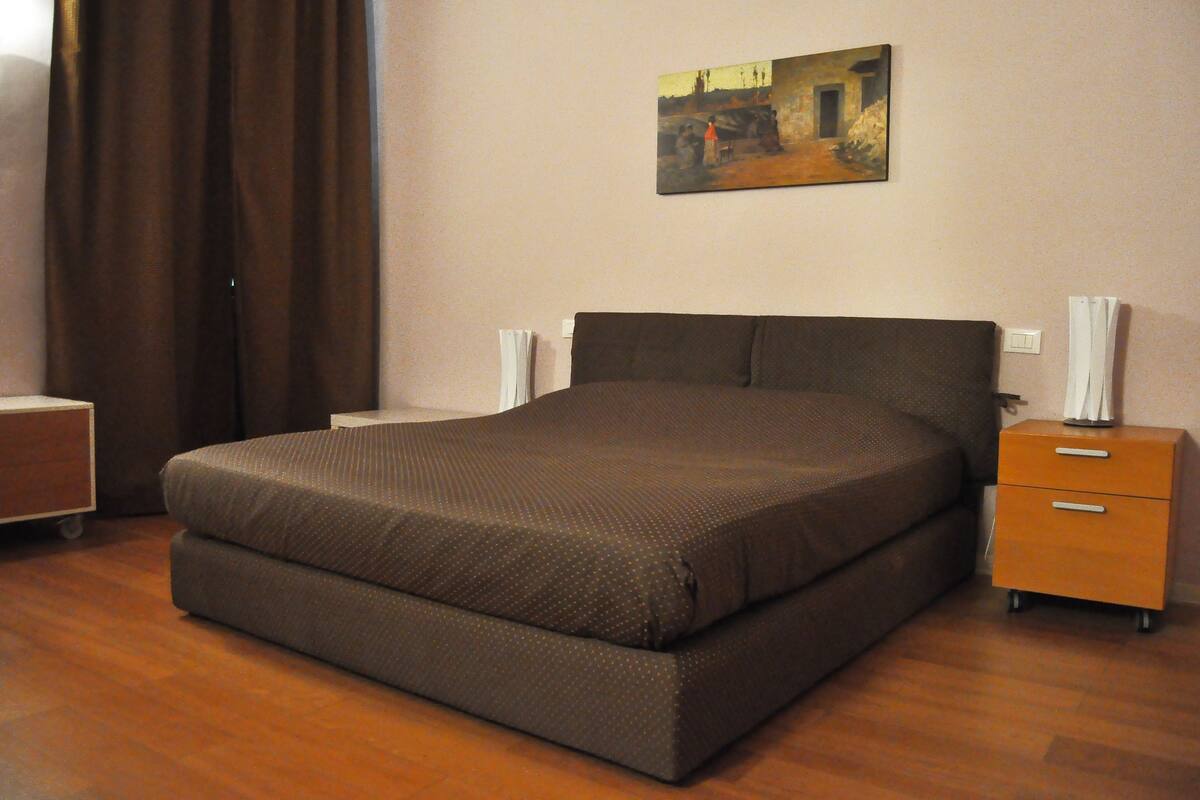 Een netjes opgemaakt bed in een kamer met een houten vloer, een nachtkastje met een lamp en een schilderij aan de muur.