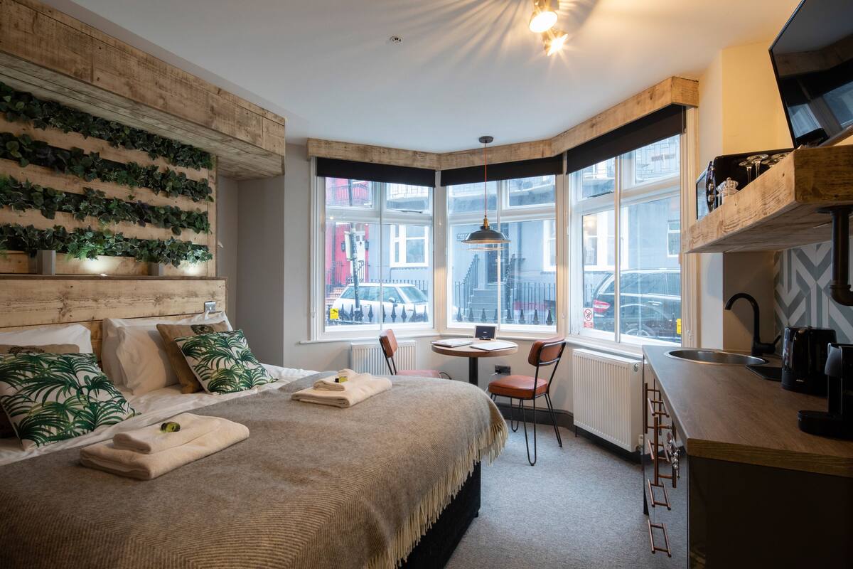A hotel room with a bed, a desk, a chair, a window with a view of the city, and a kitchen area.
