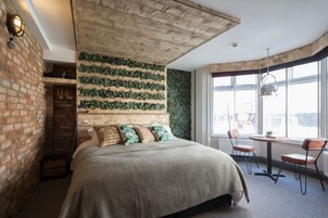 Un dormitorio con pared de ladrillo, cabecera de madera, cama con ropa de cama verde y beige, una mesita con sillas y un ventanal con vistas.