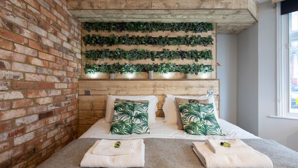 Una habitación con pared de ladrillo, techo de madera y cama con almohadillas estampadas en verde.