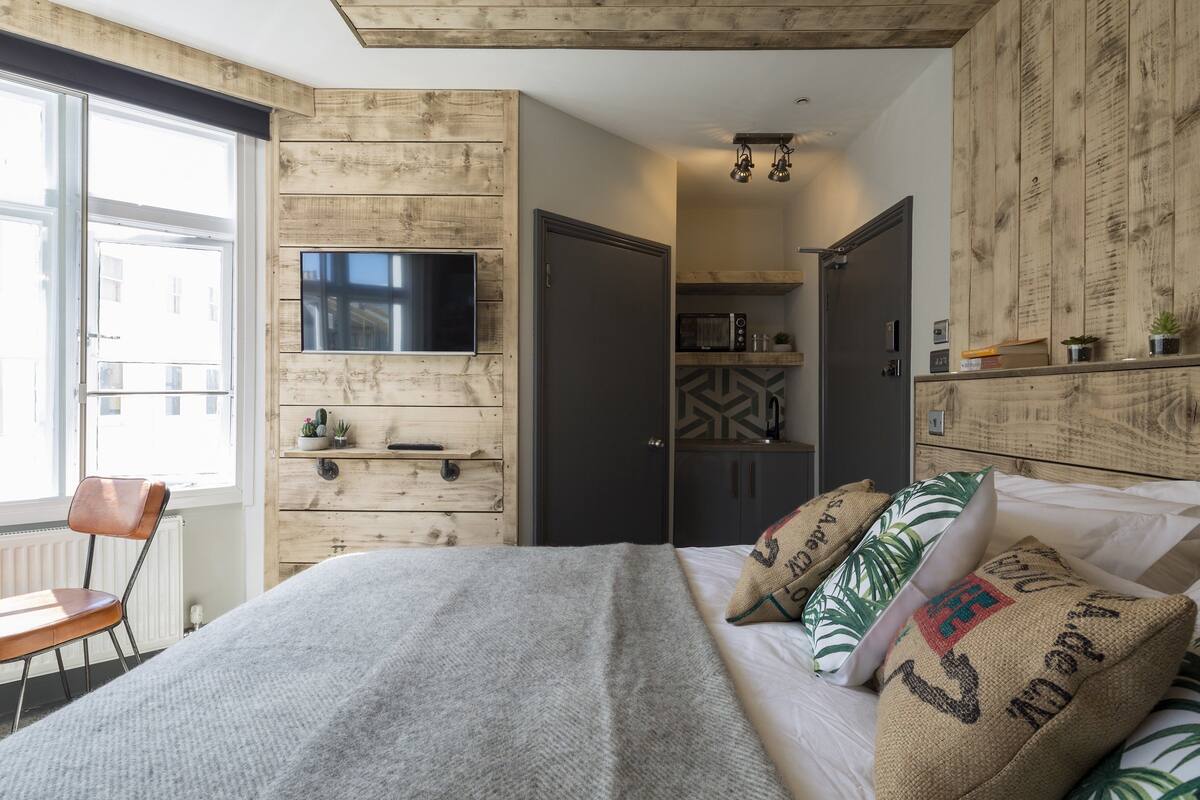 A bedroom with a bed, a TV mounted on the wall, a window, and a door leading to a kitchen area.