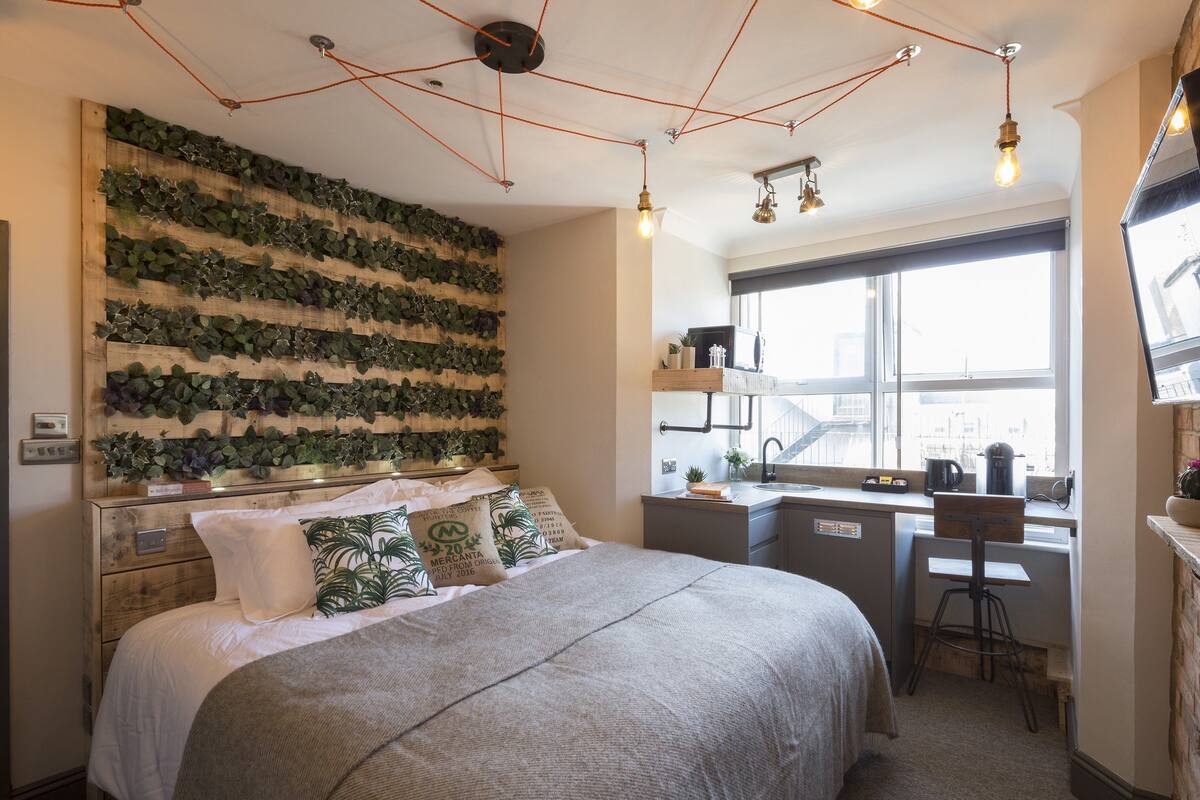 A bedroom with a bed, a desk, a chair, a window, and a wall-mounted plant display.