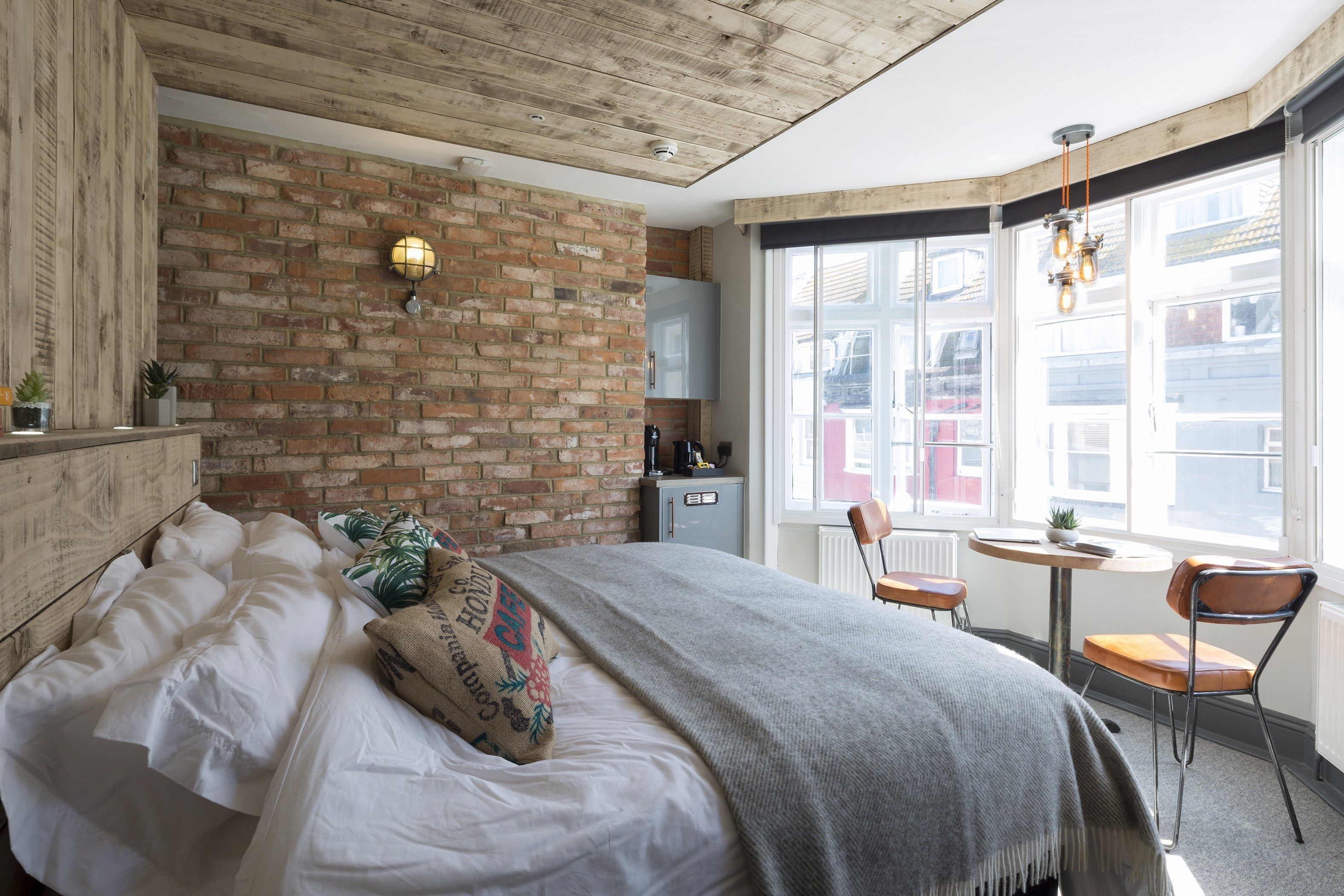 Een slaapkamer met een bakstenen muur, een bed met witte lakens, een grijze plaid, een klein rond tafeltje met een stoel en een raam met uitzicht op gebouwen.