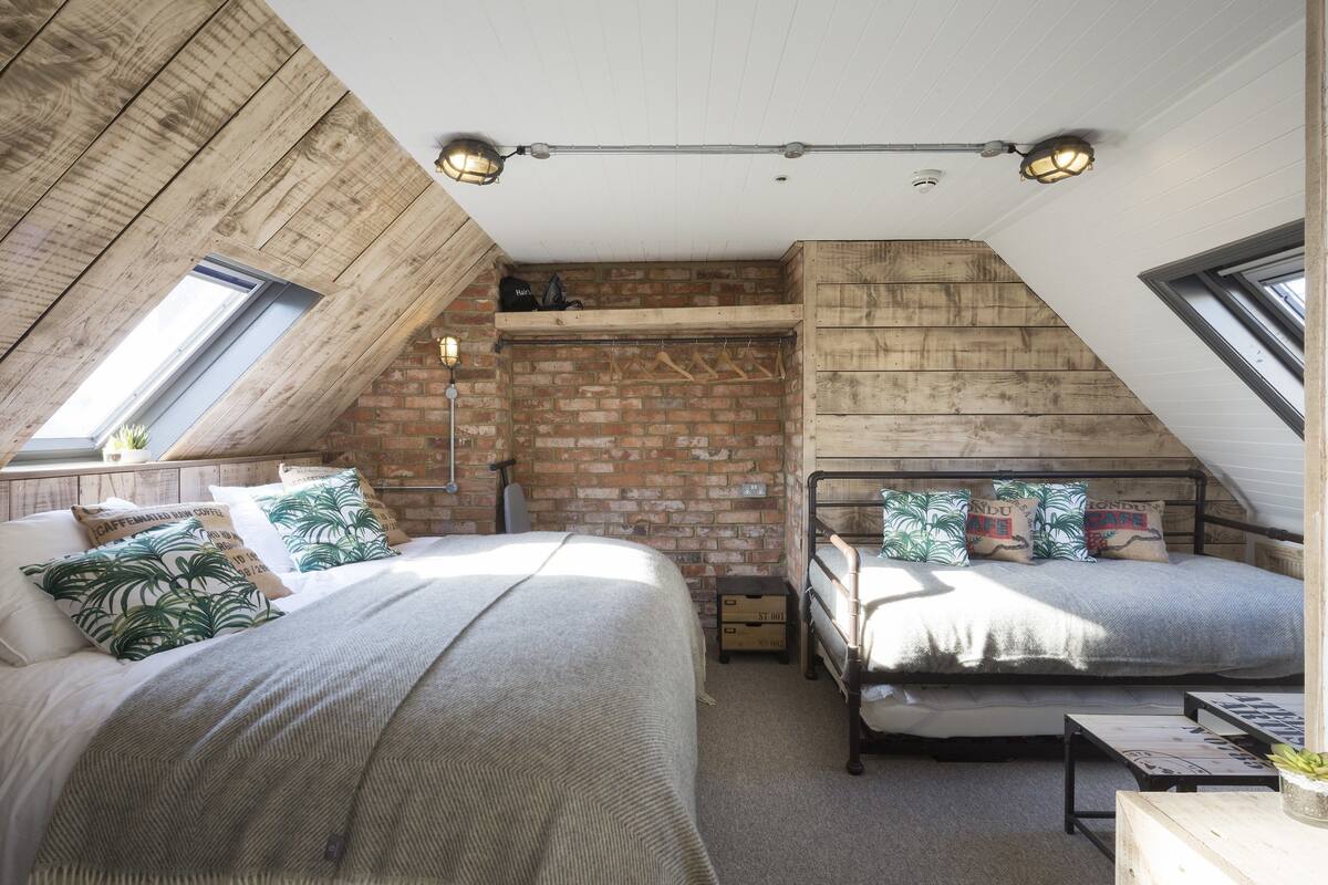 A loft bedroom with two beds, a brick wall, wooden beams, and skylights.