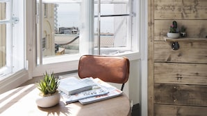 Una habitación con pared de madera, una ventana con persianas blancas, una planta en maceta, una silla y una mesa con revistas.