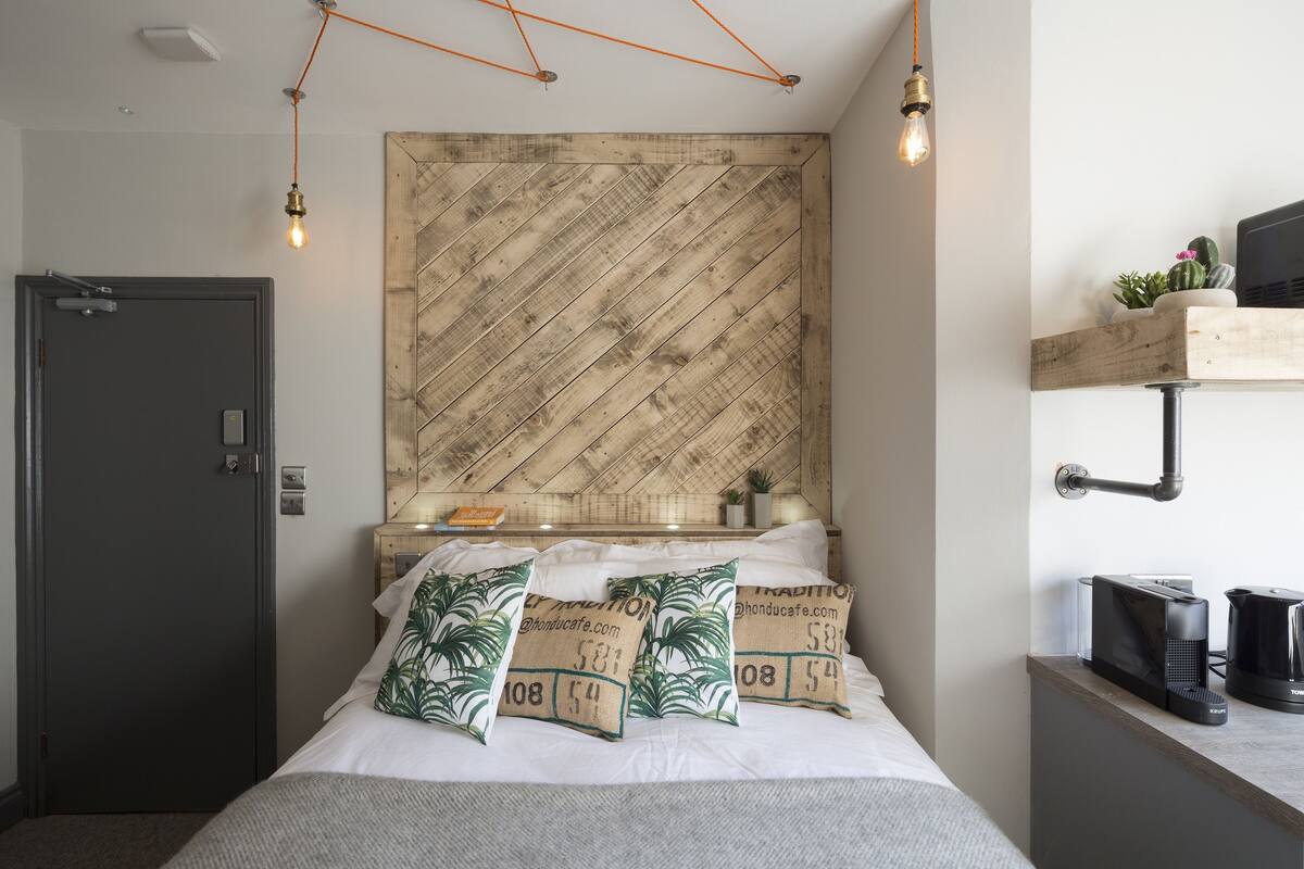 A modern hotel room with a wooden headboard, a bed with white linens and green patterned pillows, a bedside table with a coffee maker, and a mounted TV.