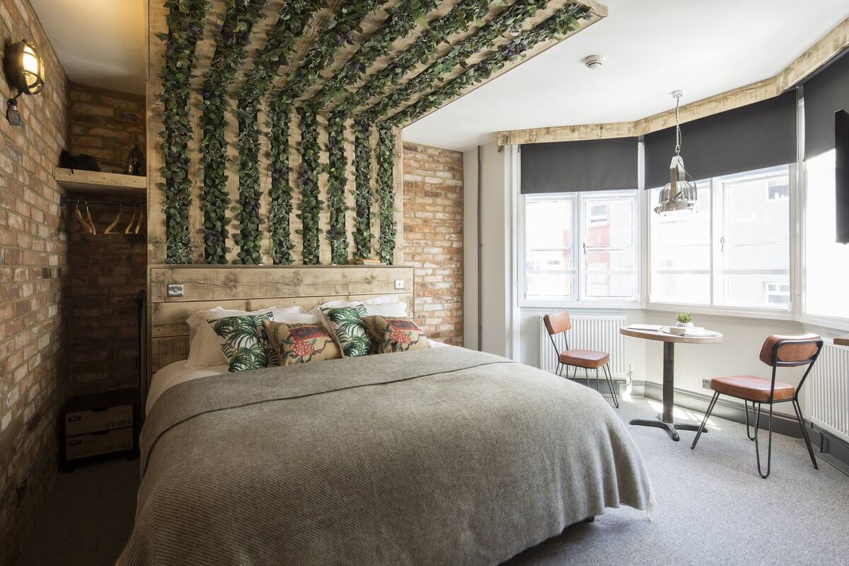 A bedroom with a brick wall, a wooden headboard, a bed with a grey blanket, and a window with a view of the outside.