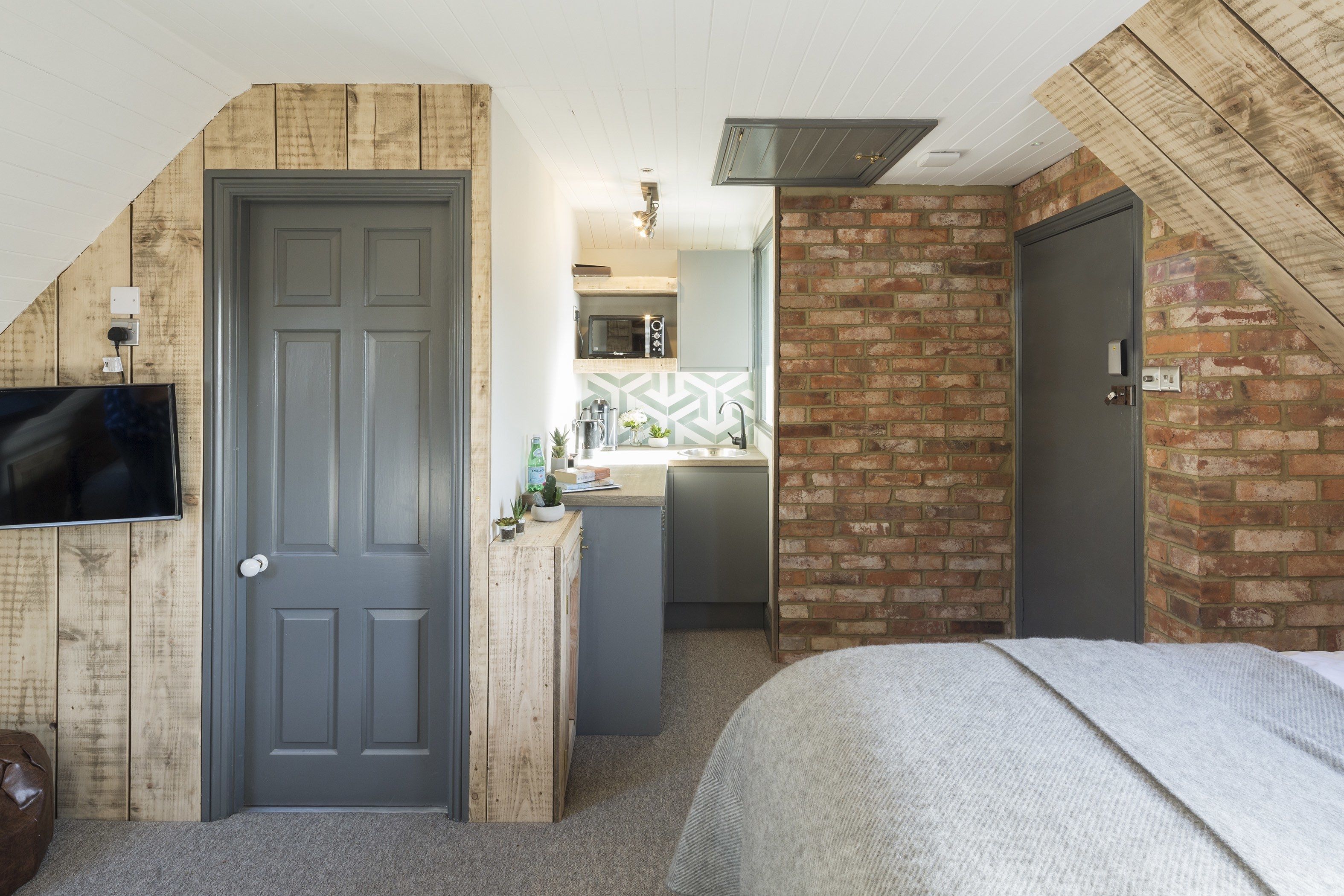 A bedroom with a bed, a TV mounted on the wall, a wooden door, and a brick wall.