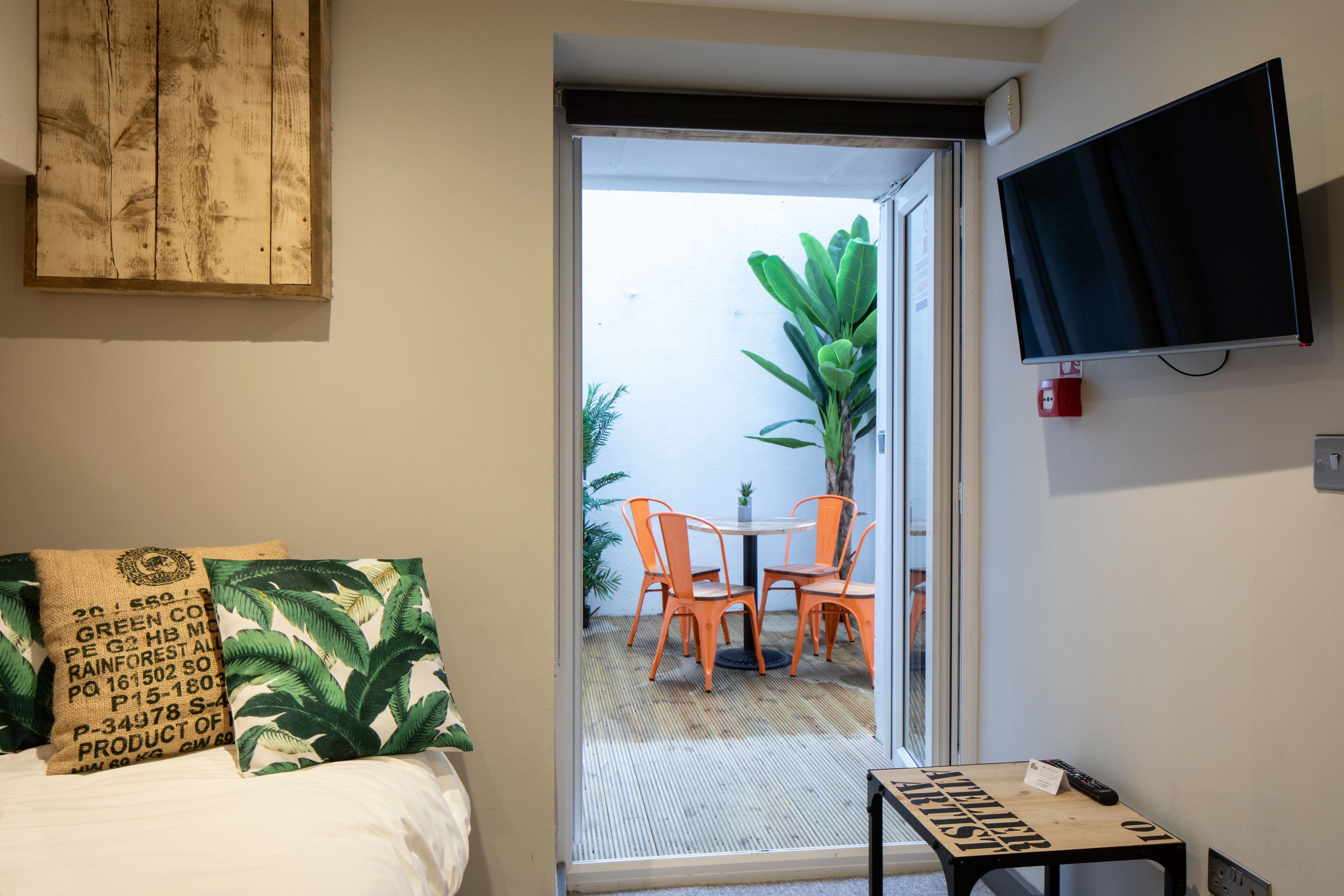 A room with a bed, a TV mounted on the wall, a small table with a remote, and a sliding glass door leading to an outdoor area with a table and chairs.