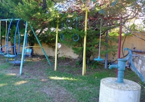 Kids can play on the swing set and climbing structure in this outdoor play area surrounded by a low wall and lush greenery.