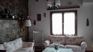 A rustic living room with a stone wall, wooden ceiling beams, a floral sofa, a wooden coffee table, and a large window with curtains. - Villa Eva Agios Nikolaos (Sithonia)