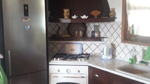 A compact kitchen with a refrigerator, oven, and microwave. There is a clock on the wall and a window with a view. - Villa Eva Agios Nikolaos (Sithonia)