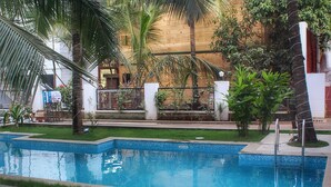 A swimming pool with a metal ladder, surrounded by palm trees and a building with a wooden facade. - Mariners Bay Resorts (Mandrem)
