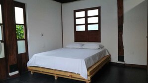 Un dormitorio con una cama de madera, ropa de cama blanca, un ventanal con múltiples vidrios y un suelo de baldosas oscuras.