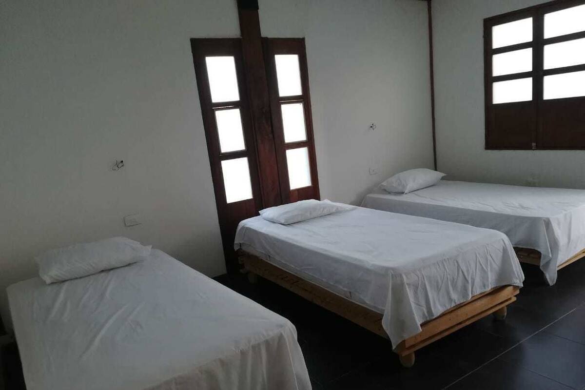 Ein Zimmer mit drei an einer Wand angeordneten Einzelbetten, einem großen Fenster mit zahlreichen Sprossen und einer Tür mit Holzrahmen.