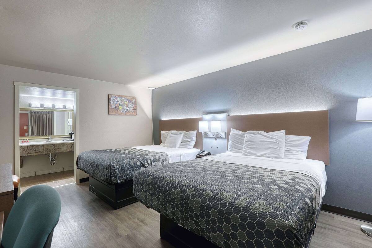 A hotel room with two beds, a desk with a chair, a bathroom with a sink and mirror, and a small artwork on the wall.
