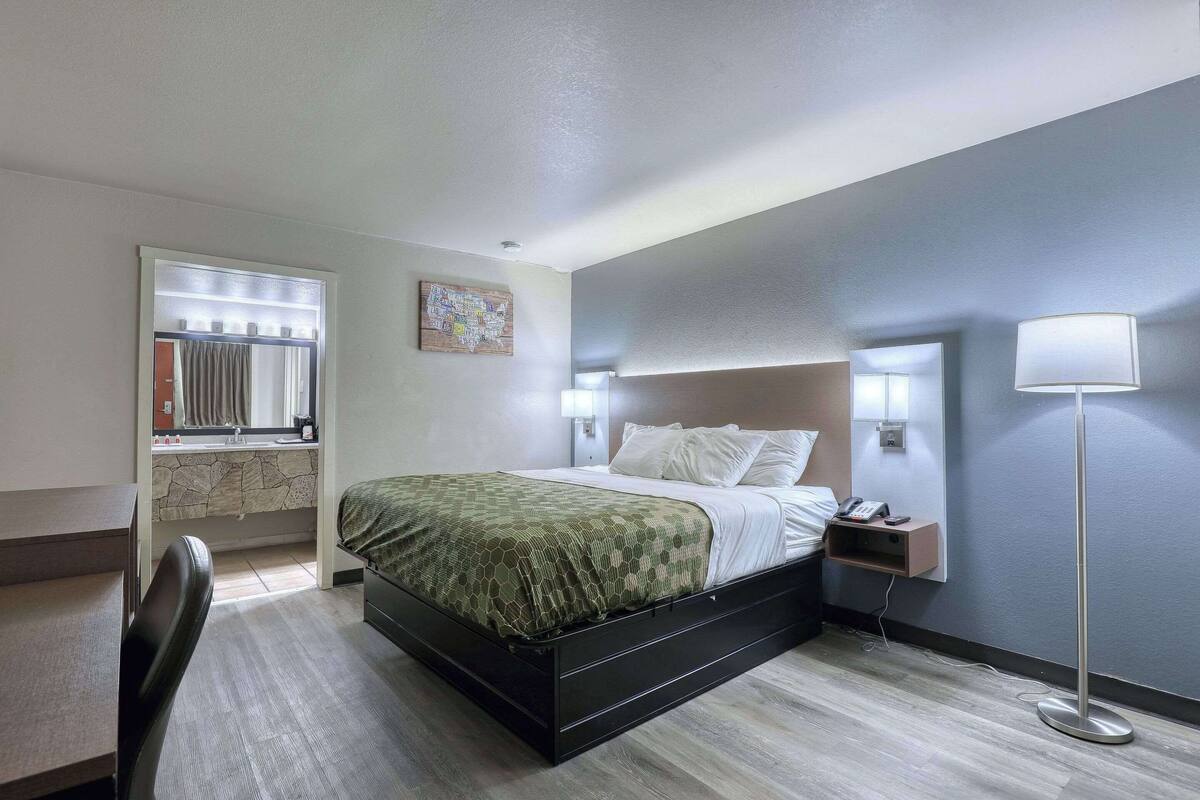 A hotel room with a bed, bedside table, lamp, and a small desk with a chair.