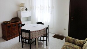 A dining area with a round table and four chairs, a wooden cabinet, a white shelf, and a door. - Apartment close to the beach (Alghero)