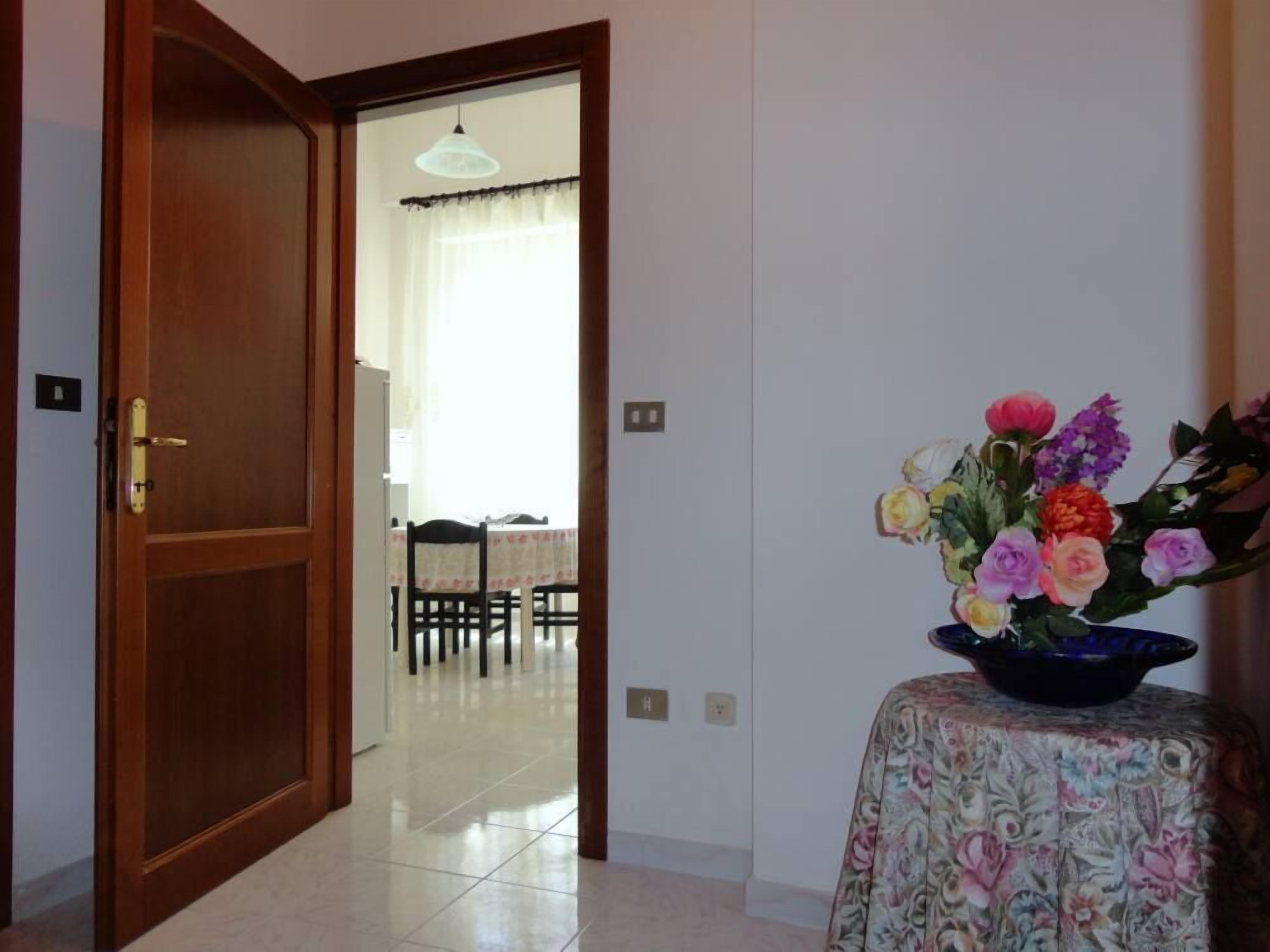 Una habitación con puerta de madera, suelo de baldosas, una mesa con sillas y mantel con motivos florales.
