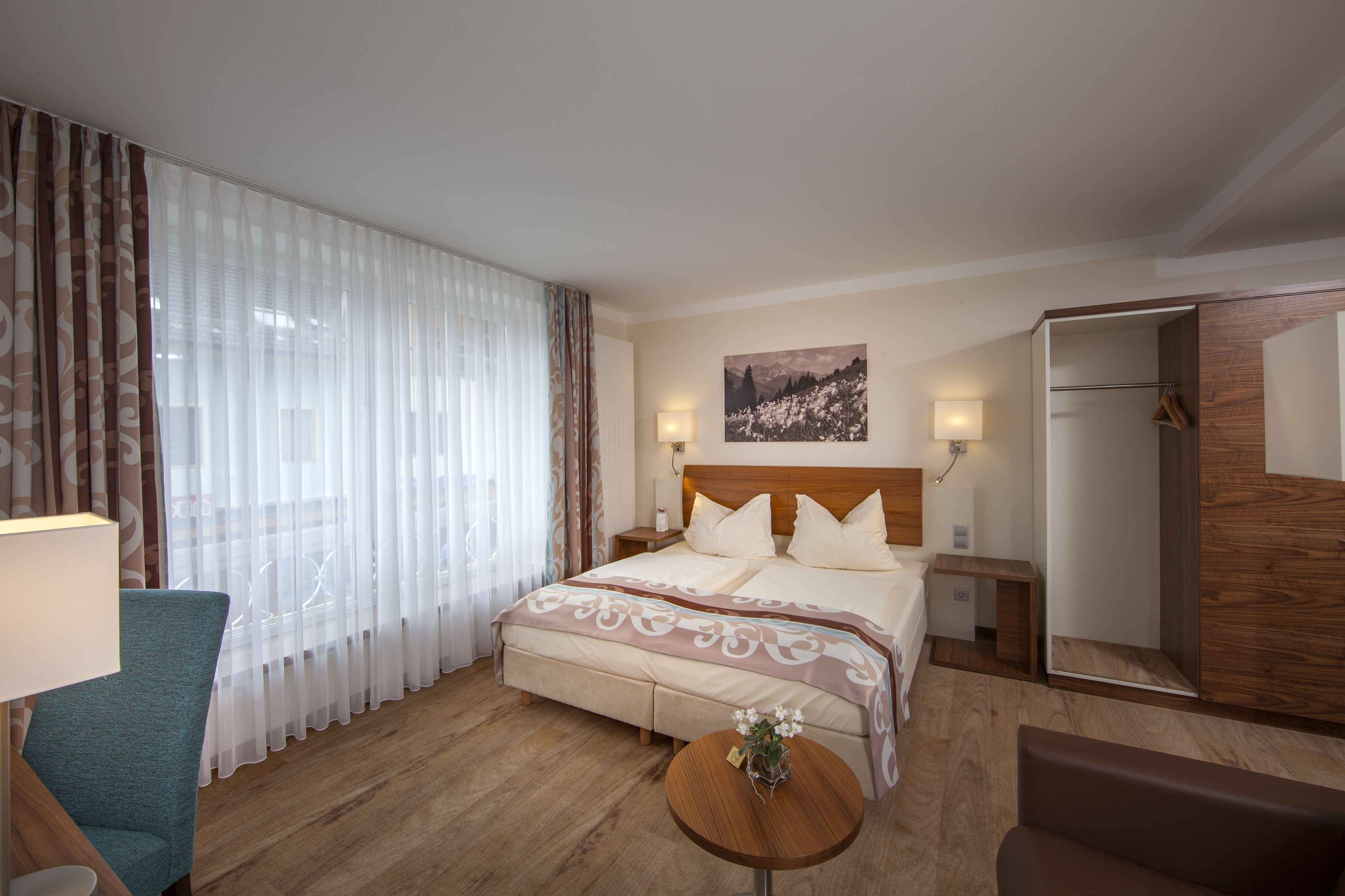 Quarto de hotel com uma cama grande, cabeceira de madeira, mesa de cabeceira com abajur, uma cadeira, pequena mesa redonda com um vaso e um guarda-roupa.