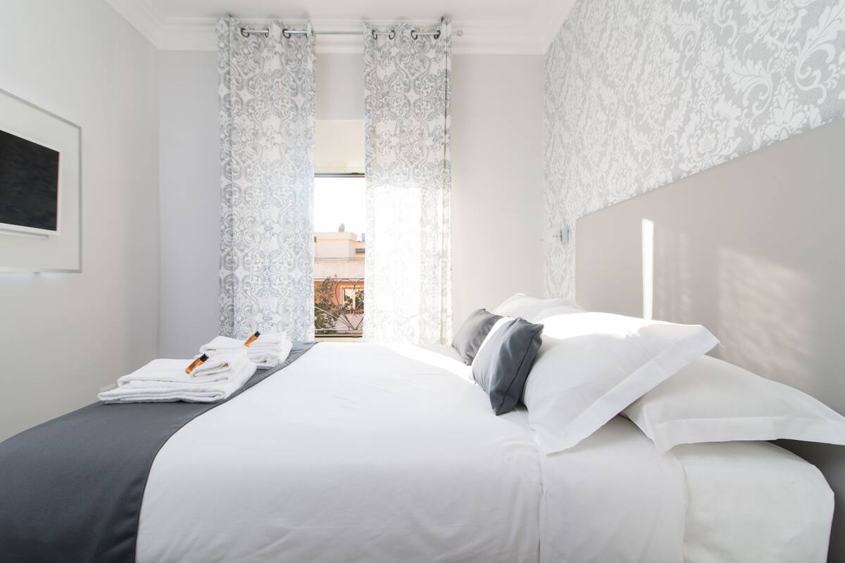 A neatly made bed with white linens and gray pillows, a window with patterned curtains, and a view of buildings outside.