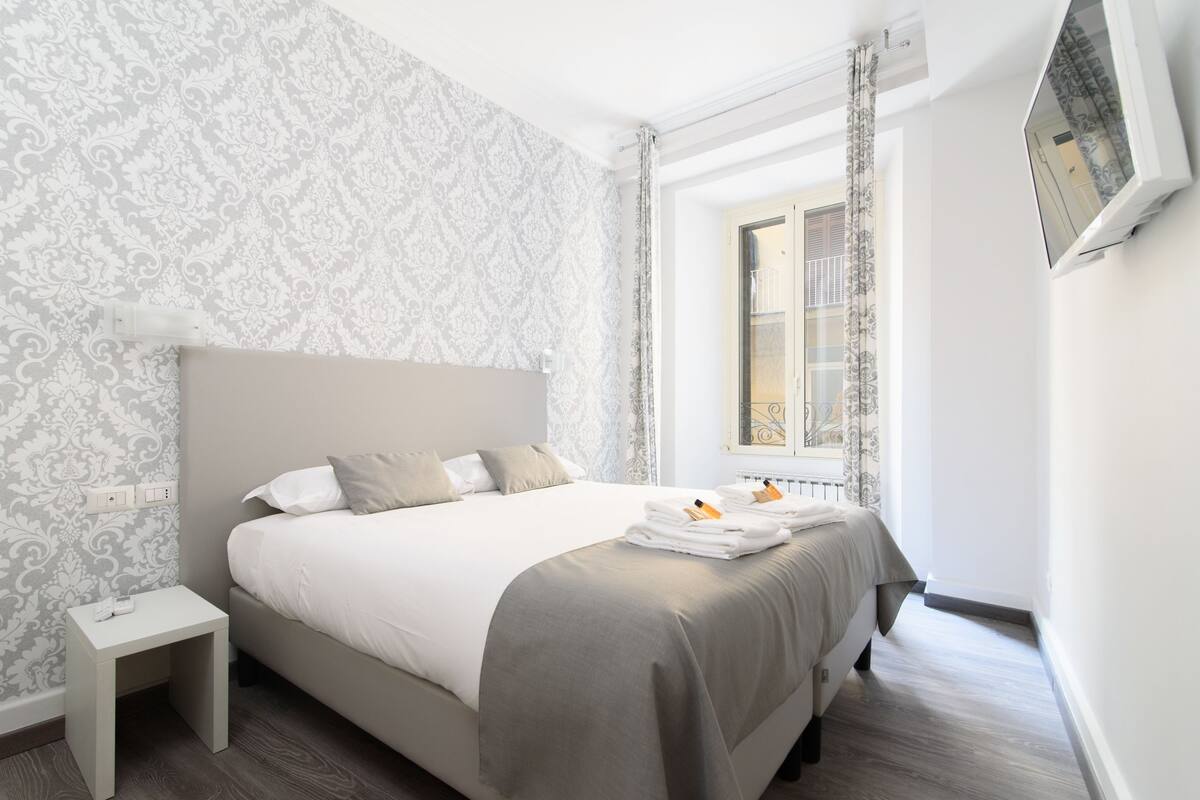 A neatly made bed with a grey headboard, a small bedside table, and a window with curtains.