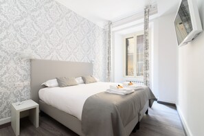 A neatly made bed with a grey headboard, a small bedside table, and a window with curtains. - White Caesar Guesthouse (Rome)