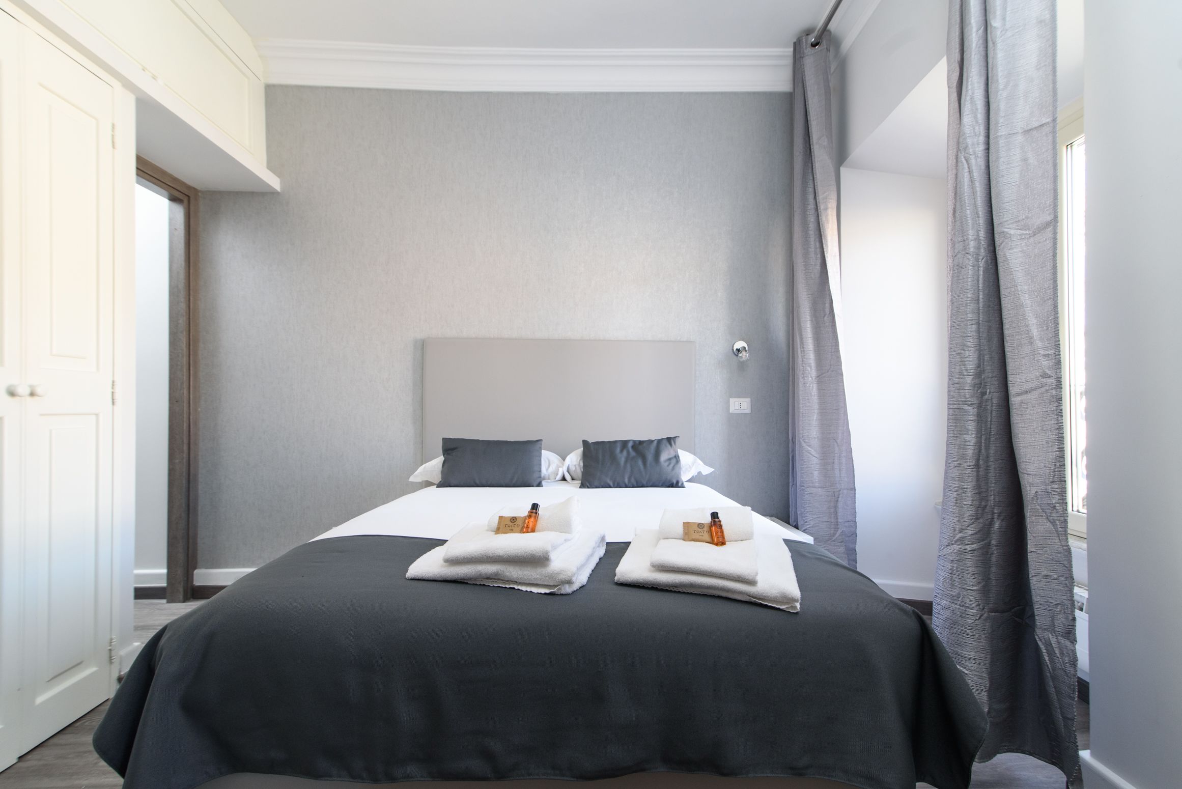 A neatly made bed with two pillows and two folded towels, a grey headboard, and a window with curtains.