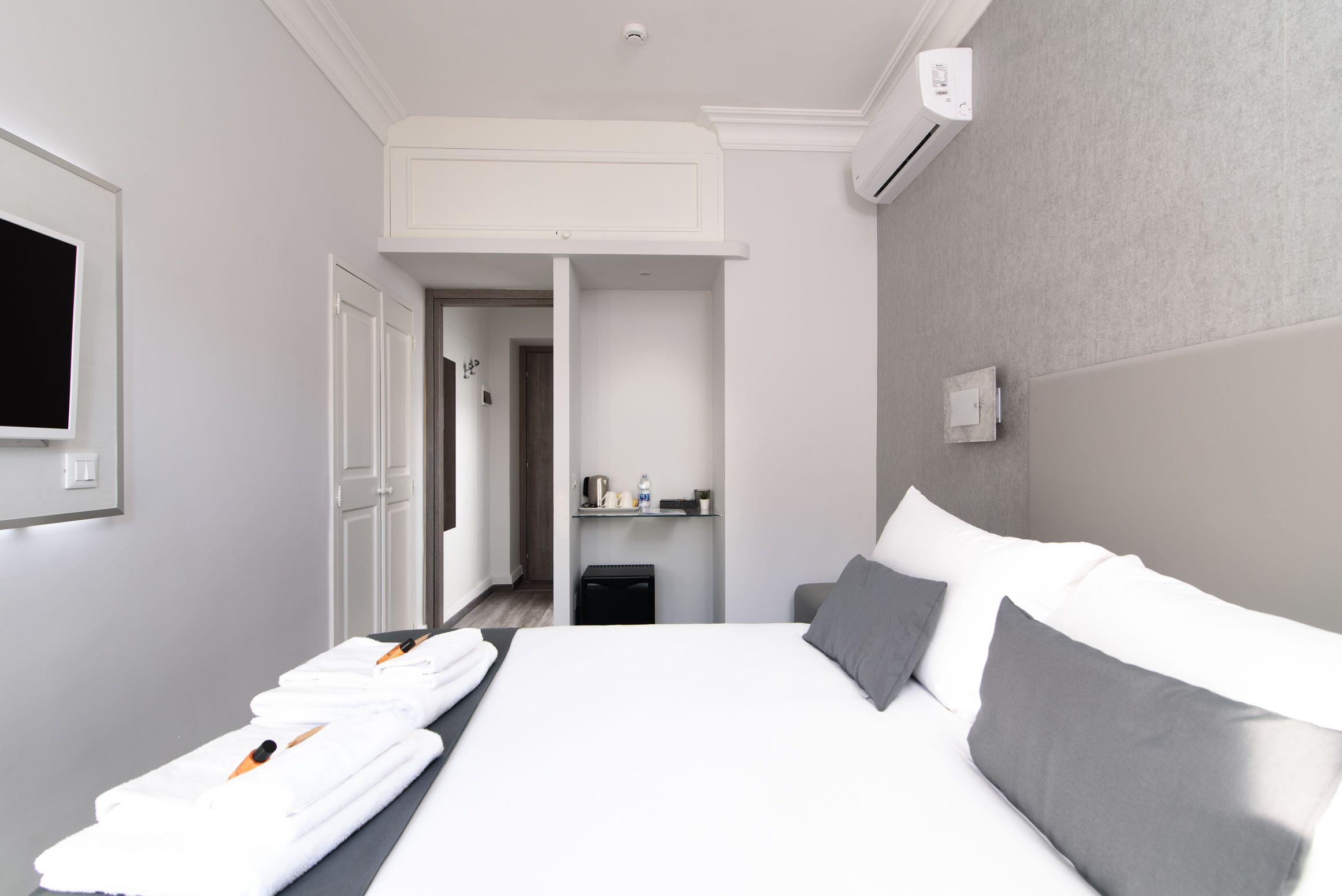 A hotel room with a neatly made bed, a small bedside table, a wall-mounted TV, and a door leading to another room.