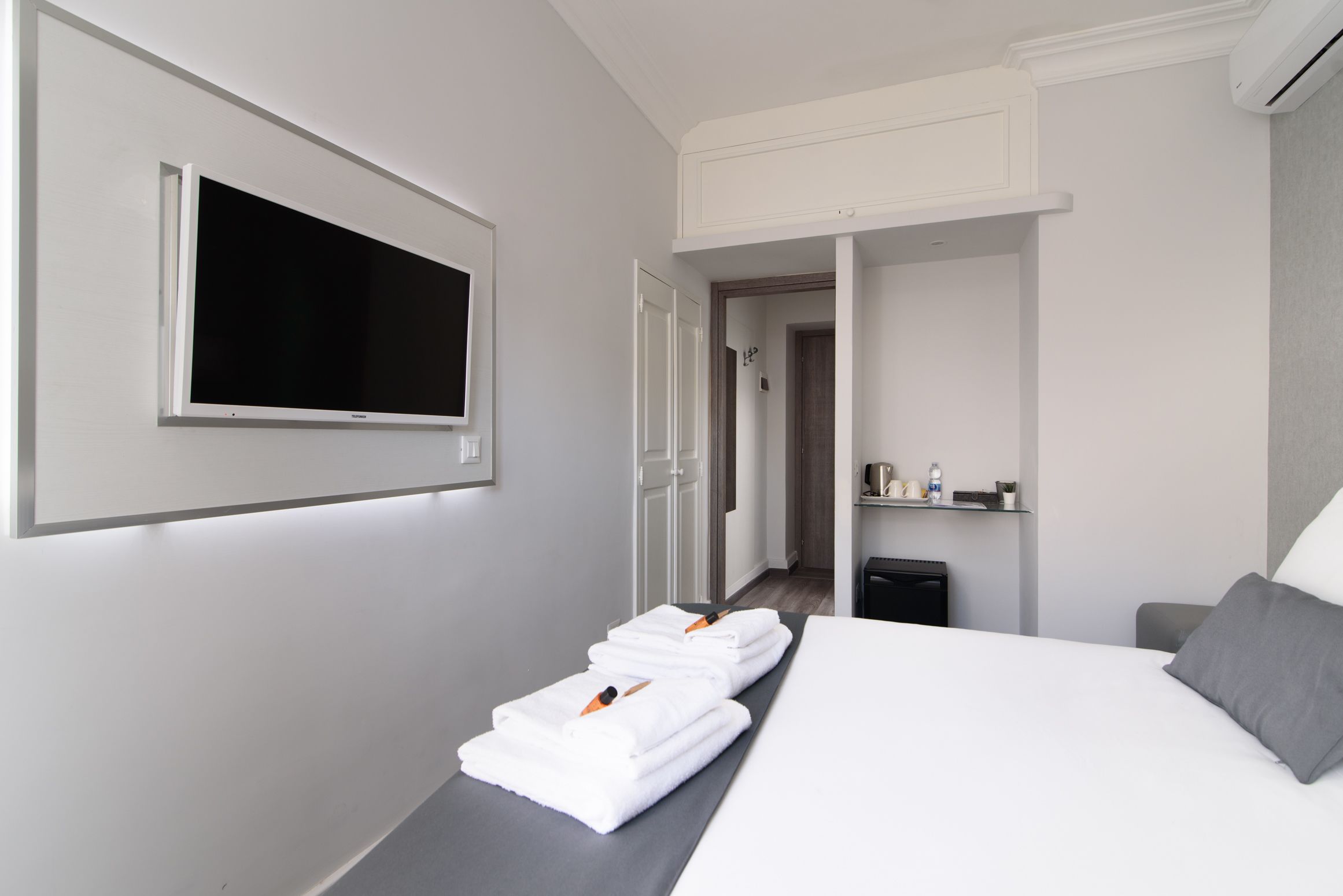 A hotel room with a bed, a TV mounted on the wall, a towel rack with towels, a bedside table, and a small shelf with toiletries.