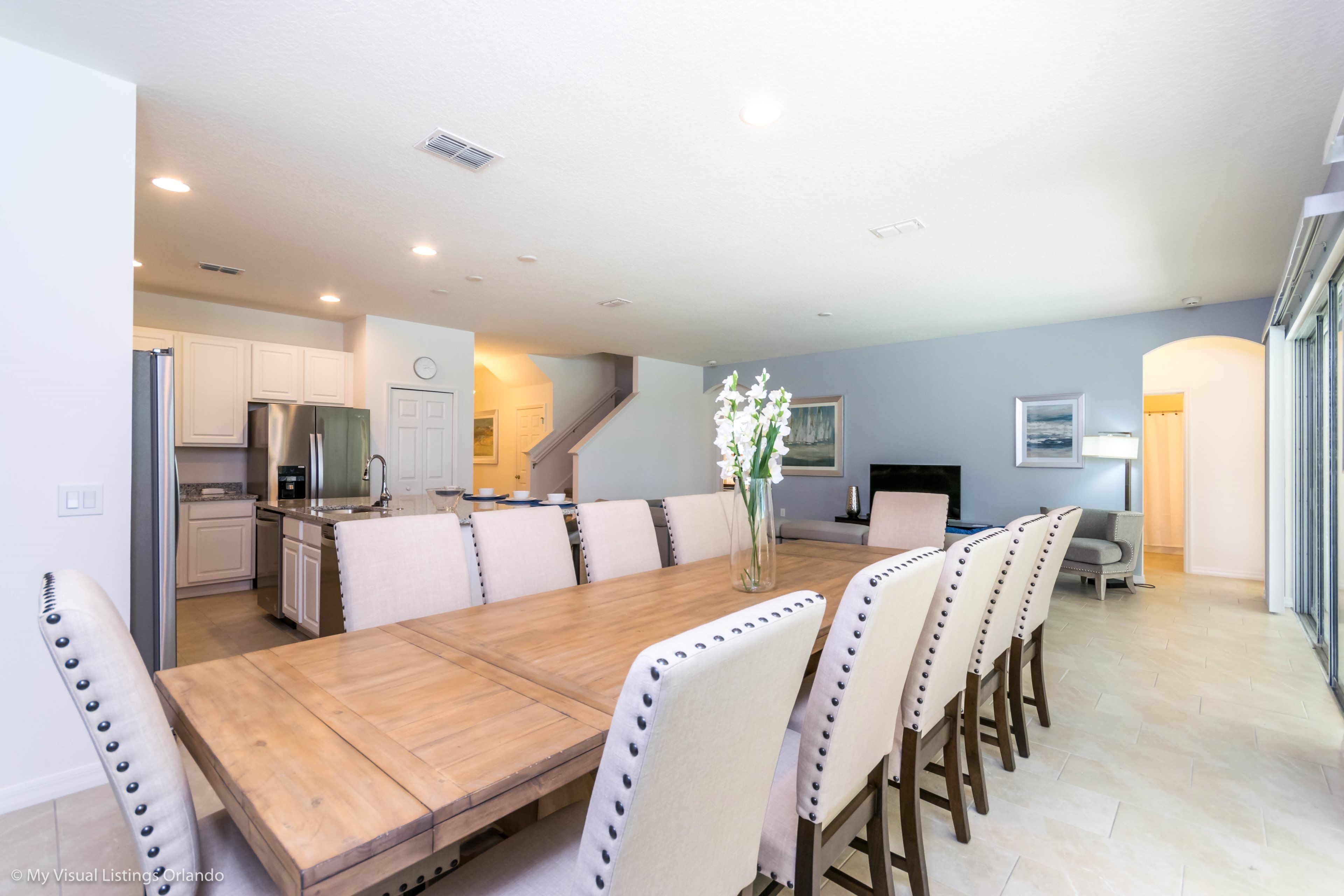 A spacious dining area with a long wooden table and multiple chairs, a kitchen in the background, and a staircase leading to another level.