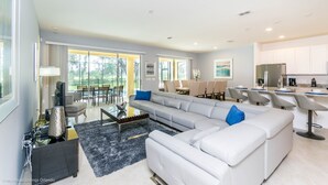 A modern living room with a sectional sofa, a coffee table, and a TV. There is a dining area with chairs and a table. The kitchen has white cabinets and a refrigerator. - H6108amazing Brand new 9 Bedroom 7 Bathroom Resort (Davenport)