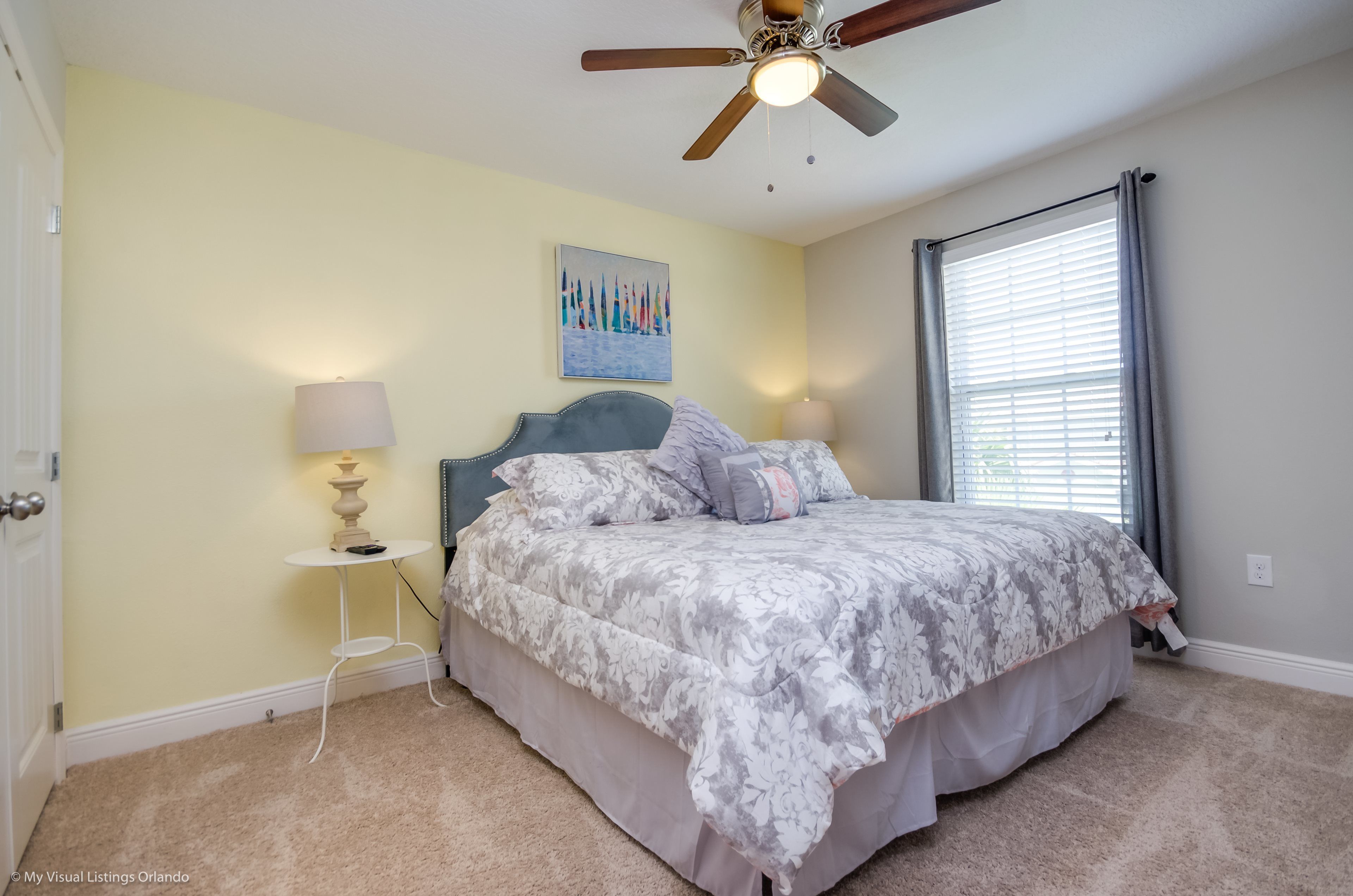 A bedroom with a bed, a ceiling fan, a window with blinds, and a wall-mounted artwork.