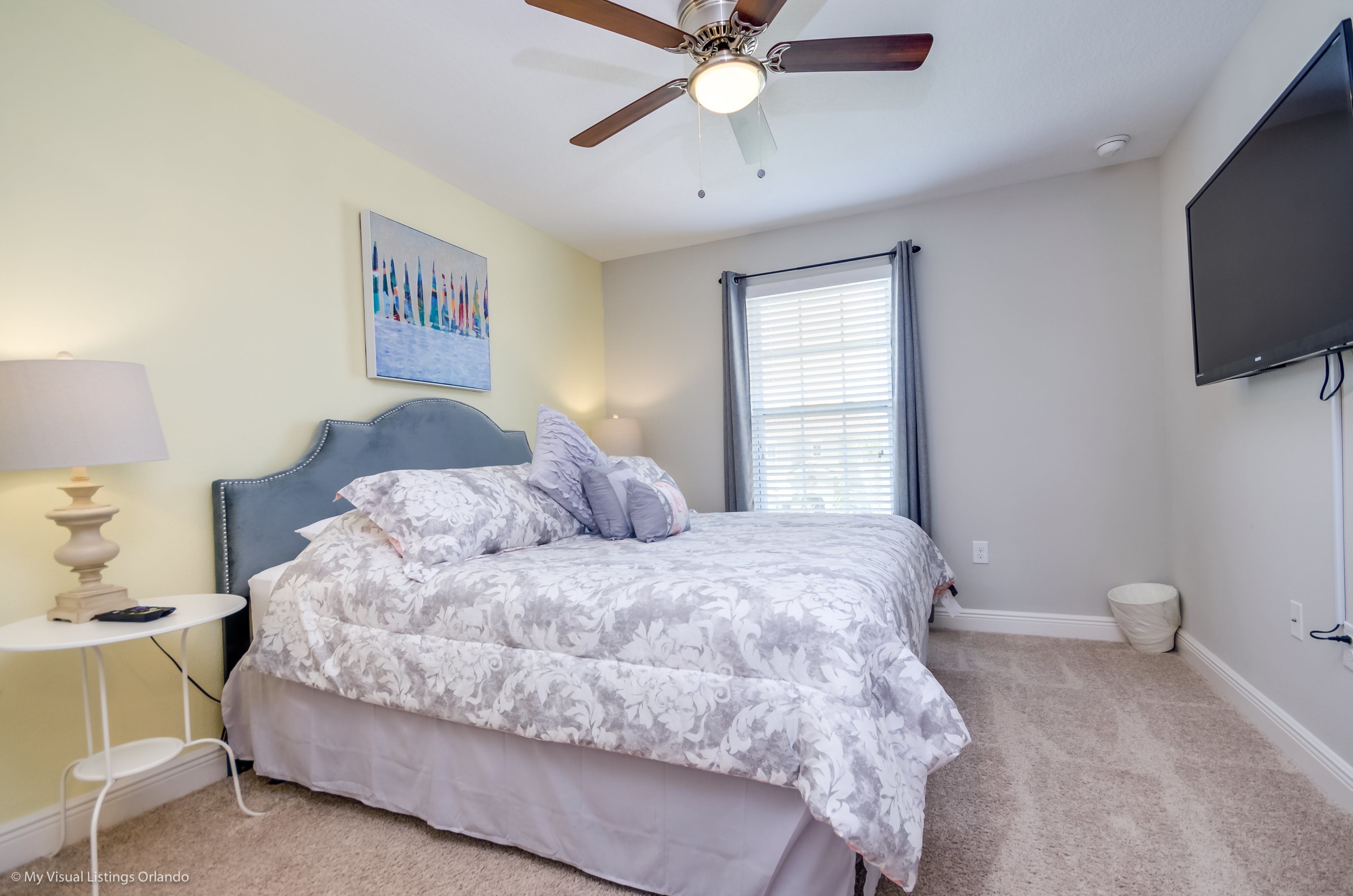 A bedroom with a bed, a ceiling fan, a TV, a lamp, and a painting on the wall.