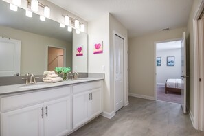 Casa de banho moderna com um lavatório grande, uma sanita dupla e um espelho com luzes. Existem duas imagens emolduradas na parede e uma porta que dá para um quarto.