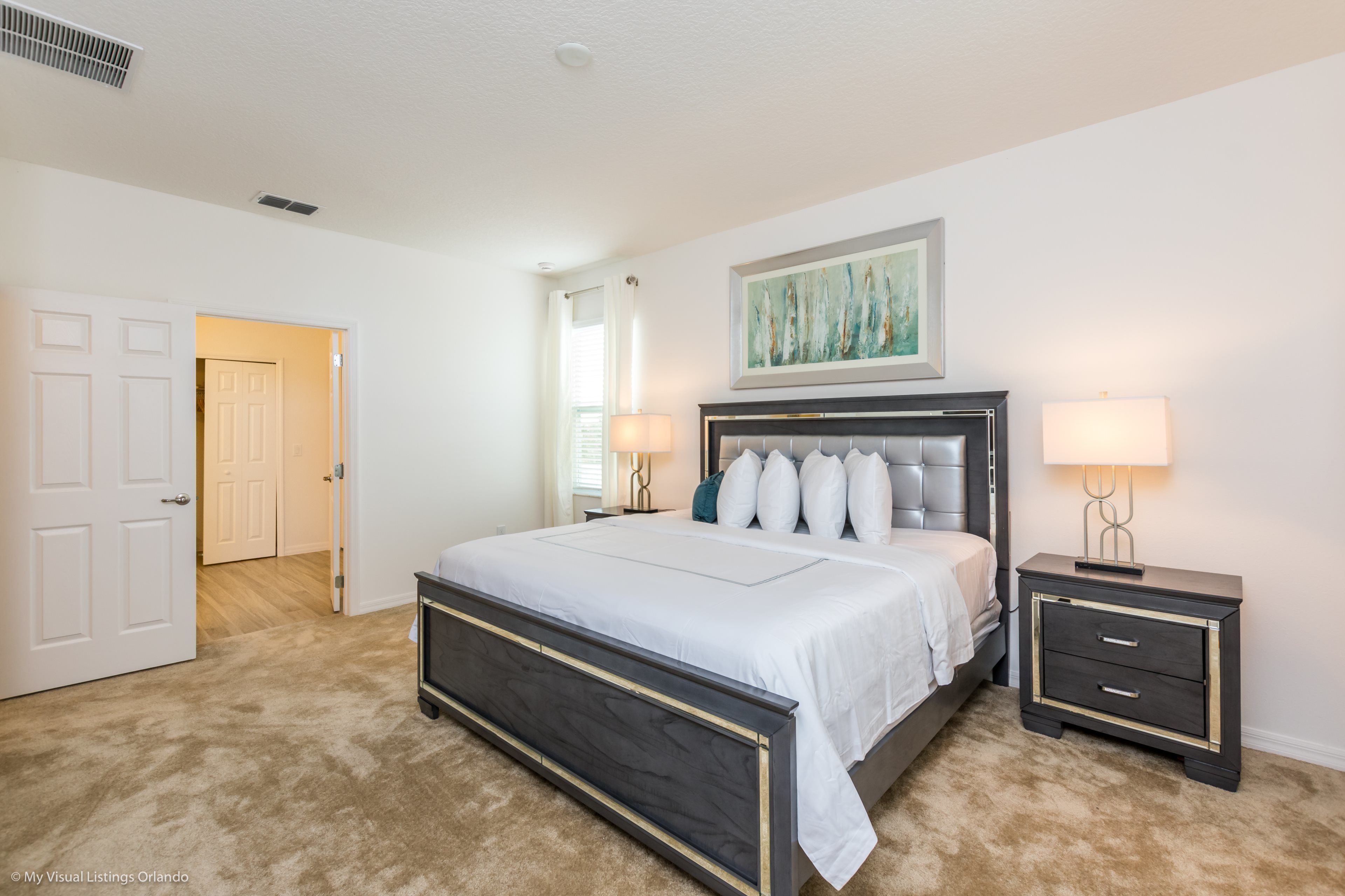 A bedroom with a large bed, two bedside tables with lamps, a door, and a ceiling vent.