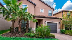 A two-story house with a beige exterior, a dark brown garage door, and a well-maintained garden with palm trees and shrubs. - H5424 Ultimate 5 Bedroom 5 Bathroom Luxury Villa (Davenport)